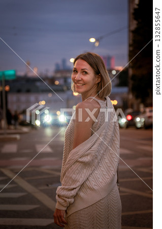 Woman in a beige knitted outfit walks through a city street at dusk, illuminated by soft streetlights and car headlights Woman in a beige knitted outfit walks through a city street at dusk, illuminated by soft streetlights and car headlights 132855647