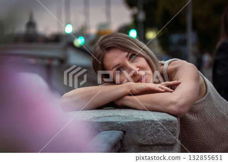 Woman in a beige knitted dress leans on a stone railing at dusk, with blurred city lights in the background Woman in a beige knitted dress leans on a stone railing at dusk, with blurred city lights in the background 132855651