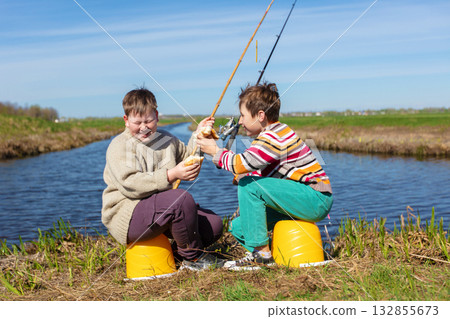 The boys are friends on a fishing trip by the river eating a bun and laughing. Children are friends on a summer fishing trip. The boys are friends on a fishing trip by the river eating a bun and laughing. Children are friends on a summer fishing trip. 132855673