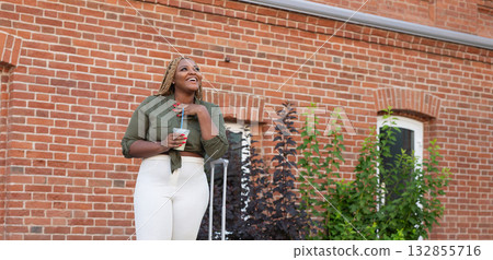 African american woman standing with coffee cup and suitcase near brick wall. Travel, lifestyle, and positive emotion in modern urban environment. Banner with copy space 132855716