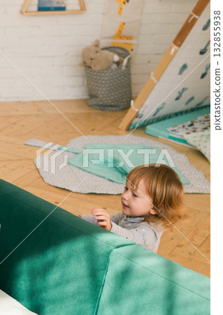 Happy little girl smiling and playing behind green sofa in cozy children room. Joy, curiosity, and early development at home. Happy little girl smiling and playing behind green sofa in cozy children room. Joy, curiosity, and early development at home. 132855938