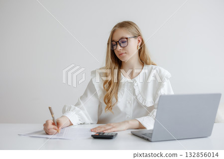 Young woman working at desk with laptop and calculator while taking notes in a bright, modern workspace during the day 132856014