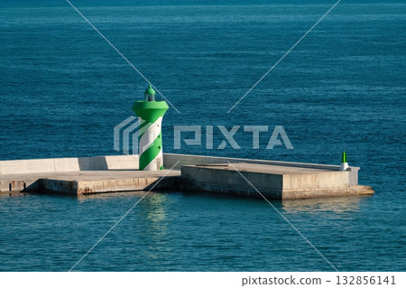 Green lighthouse with a white spiral stripe stands on a concrete breakwater in the port 132856141