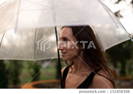 Woman, umbrella, rain, protects herself outdoors with a clear umbrella on a wet day, smiling gently Woman, umbrella, rain, protects herself outdoors with a clear umbrella on a wet day, smiling gently 132856306