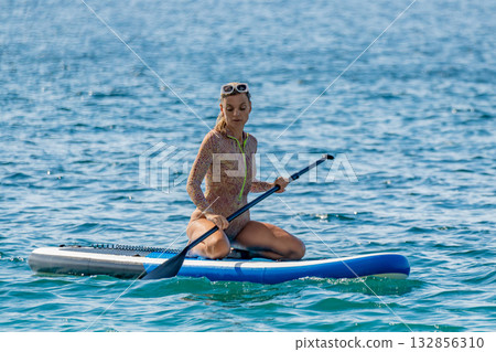 Paddleboarding woman ocean, active female kneeling on a stand up paddleboard in tranquil blue sea. Paddleboarding woman ocean, active female kneeling on a stand up paddleboard in tranquil blue sea. 132856310