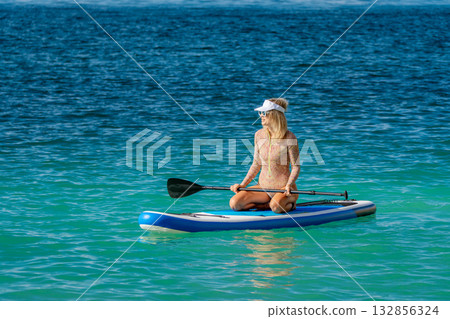 Woman, paddleboard, ocean, young woman kneeling on a stand-up paddleboard enjoying summer water sports in clear blue water with copy space. 132856324