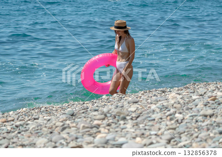 Woman Beach Sea, young woman in white bikini, pink ring, exits water onto pebble beach, summer. Woman Beach Sea, young woman in white bikini, pink ring, exits water onto pebble beach, summer. 132856378