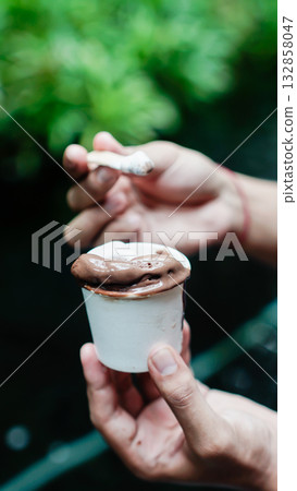 Chocolate Artisan Gelato in Paper Cup Held by Hand, Close-Up Dessert with Spoon and Natural Green Background 132858047