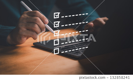 Person using a pen with a laptop to tick off items on a virtual checklist, symbolizing productivity, workflow, project planning and efficiency tools. Digital checklist and task management concept. 132858241