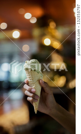 Close up of hand holding a cone of pistachio ice cream with blurred bokeh background in warm cafe lighting, showing creamy texture and cozy dessert atmosphere for lifestyle and food concept Close up of hand holding a cone of pistachio ice cream with blurred bokeh background in warm cafe lighting, showing creamy texture and cozy dessert atmosphere for lifestyle and food concept 132858797