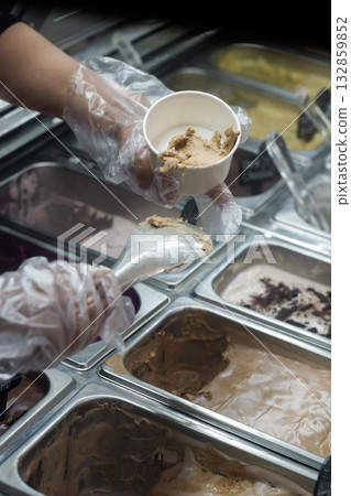 Scooping creamy gelato into a white cup at ice cream shop counter, close up of hand wearing plastic gloves, professional dessert preparation at cafe with various ice cream flavors in display freezer 132859852