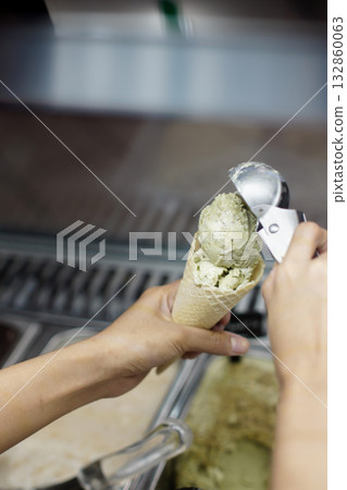 Barista scooping creamy green tea gelato into waffle cone at ice cream shop counter, close up of hand using metal scoop, fresh frozen dessert preparation with soft lighting and clean background 132860063