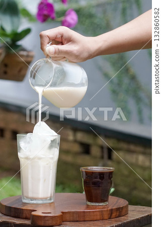 Hand pouring fresh milk into glass with ice to prepare iced latte, cold coffee drink on wooden board with espresso shot, outdoor cafe scene, refreshing summer beverage lifestyle concept 132860582