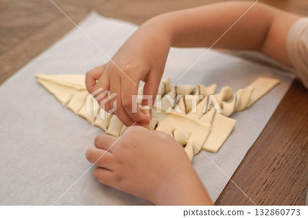 Close-up of child shaping Christmas tree-shaped pastry dough on parchment paper. Concept of holiday baking, family bonding, and creative learning at home. Close-up of child shaping Christmas tree-shaped pastry dough on parchment paper. Concept of holiday baking, family bonding, and creative learning at home. 132860773