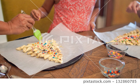 Family decorating Christmas tree-shaped pastry with colorful toppings and egg wash during a festive baking activity. Perfect for holiday, parenting, and family lifestyle concepts. 132860785