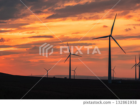 Multiple wind turbines silhouetted against a vibrant sunset sky. The turbines stand tall against the backdrop of orange and red clouds. Multiple wind turbines silhouetted against a vibrant sunset sky. The turbines stand tall against the backdrop of orange and red clouds. 132861938