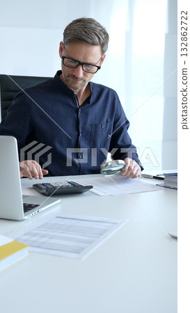 Financial analyst reviewing paperwork, using calculator and magnifying glass while examining corporate budget reports in modern workspace. Audit and taxes, business concept Financial analyst reviewing paperwork, using calculator and magnifying glass while examining corporate budget reports in modern workspace. Audit and taxes, business concept 132862722