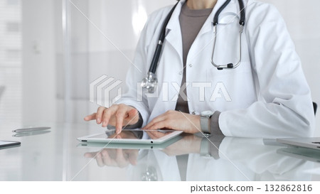 Physician in lab coat with stethoscope reviewing patient data on a tablet in a bright modern clinical office via touchscreen. Medicine and health care Physician in lab coat with stethoscope reviewing patient data on a tablet in a bright modern clinical office via touchscreen. Medicine and health care 132862816