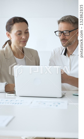 Businessman and adult businesswoman reviewing marketing analytics on laptops, working together strategically in contemporary workplace with minimalist interior design and collaborative atmosphere 132862835