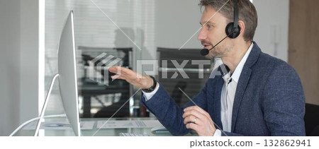 Male call center agent wearing a headset, providing online customer support and consulting clients during a video call. Business communication concept 132862941