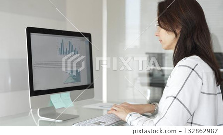 Businesswoman at glass office desk analyzing charts and graphs on computer screen, reviewing financial reports and performance data for planning. Business people concept 132862987