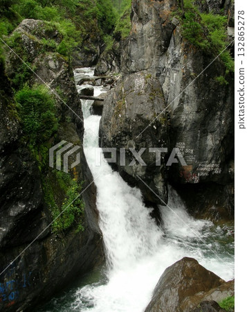 A turbulent stream of clear water, a waterfall flows down from high mountains down over rocks between high mountains, which are covered with green grass. 132865278