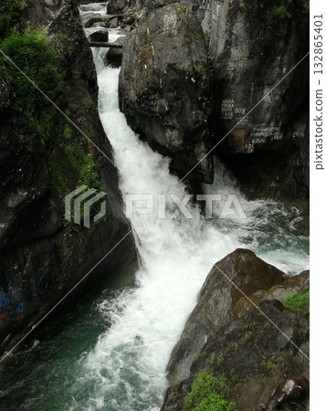 A turbulent stream of clear water, a waterfall flows down from high mountains down over rocks between high mountains, which are covered with green grass A turbulent stream of clear water, a waterfall flows down from high mountains down over rocks between high mountains, which are covered with green grass 132865401