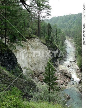 A turbulent stream  water, a waterfall flows down from high mountains down over rocks between high mountains, which are covered with green grass and trees. 132865508