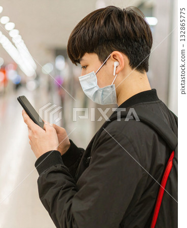 A young man wearing a mask and earphones and operating a smartphone 132865775