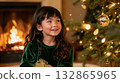Smiling girl in green velvet dress sitting near a glowing Christmas tree and cozy fireplace, enjoying holiday warmth and magic.  132865965