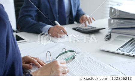 Business colleagues are working together, analyzing financial documents with a magnifying glass, calculating and taking notes in the office. Audit and taxes in business 132866218