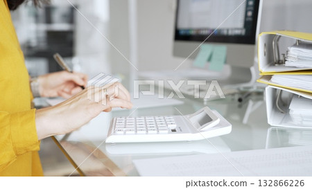 Woman in yellow blouse calculating finances at office desk, using calculator and writing, surrounded by documents and binders. Audit and taxes in business Woman in yellow blouse calculating finances at office desk, using calculator and writing, surrounded by documents and binders. Audit and taxes in business 132866226