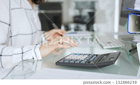 Woman calculating finances with black calculator, managing taxes, invoices, and accounting documents in office. Audit and taxes in business 132866239