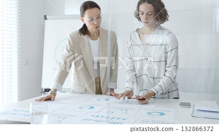 Professional businesswomen analyzing financial charts, collaborating in sleek corporate workspace, highlighting data. Business people concept 132866372