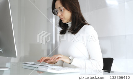 Woman wearing white casual sweater and glasses typing on computer keyboard in modern office, working on business project, sending emails, or data entry, focusing on digital tasks. Audit in business 132866410