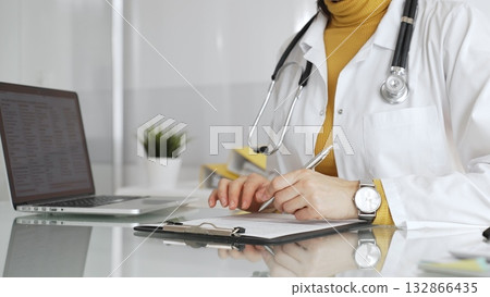 Doctor wearing stethoscope and yellow sweater under white lab coat, making notes on a clipboard in a bright medical office. Medicine, healthcare and science concept Doctor wearing stethoscope and yellow sweater under white lab coat, making notes on a clipboard in a bright medical office. Medicine, healthcare and science concept 132866435