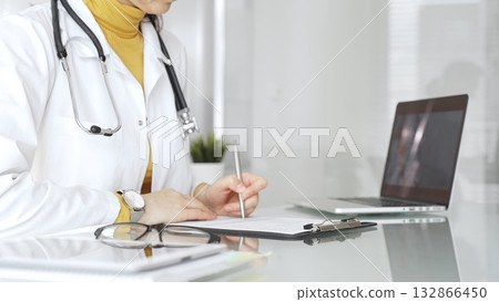Doctor wearing stethoscope and yellow sweater under white lab coat, making notes on a clipboard in a bright medical office. Medicine, healthcare and science concept 132866450