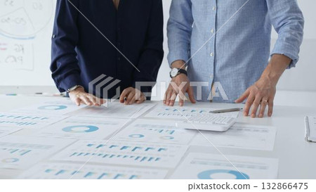 Focused business professionals analyzing financial charts and graphs, discussing market trends and investment strategies while leaning over white desk surface. Focused business professionals analyzing financial charts and graphs, discussing market trends and investment strategies while leaning over white desk surface. 132866475