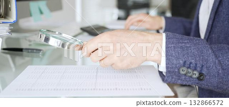 Male accountant holding magnifying glass, checking financial documents on office desk, analyzing taxes and data. Audit and taxes in business Male accountant holding magnifying glass, checking financial documents on office desk, analyzing taxes and data. Audit and taxes in business 132866572