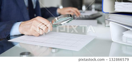 Hand holding magnifying glass, inspecting financial document, performing audit and business analysis. Audit and taxes in business Hand holding magnifying glass, inspecting financial document, performing audit and business analysis. Audit and taxes in business 132866579