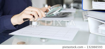 Person auditing financial document using magnifying glass on desk with binders, researching business data. Audit and taxes in business Person auditing financial document using magnifying glass on desk with binders, researching business data. Audit and taxes in business 132866593
