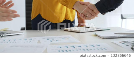 Business partners shaking hands after successful meeting in office while another person is clapping hands over financial documents, laptop and notebook 132866646