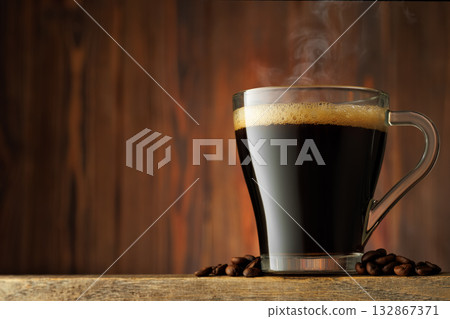 Steam rises from transparent cup of hot espresso stand on table with coffee beans scattered around it against wooden background 132867371