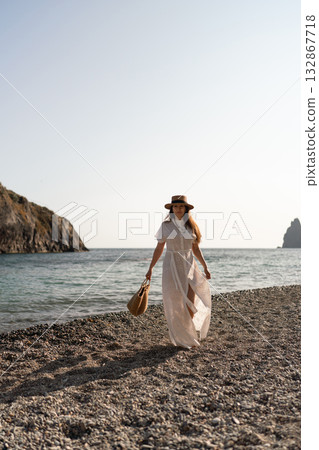 Woman, beach, vacation: elegant woman in white dress walks on pebble shore with straw bag, serene summer travel copy space Woman, beach, vacation: elegant woman in white dress walks on pebble shore with straw bag, serene summer travel copy space 132867718