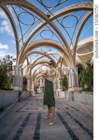 Woman Fashion Architecture - Stylish woman wearing a beige sweater and green skirt, posing under ornate arches in a modern architectural structure. Woman Fashion Architecture - Stylish woman wearing a beige sweater and green skirt, posing under ornate arches in a modern architectural structure. 132867752