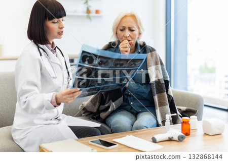 A female doctor in a white coat shows an X-ray to an elderly woman, who is coughing and wrapped in a blanket, in a medical consultation. 132868144