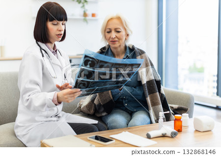 A female doctor in a white coat explains an X-ray to an elderly woman, likely discussing her diagnosis and treatment options in a home setting. A female doctor in a white coat explains an X-ray to an elderly woman, likely discussing her diagnosis and treatment options in a home setting. 132868146