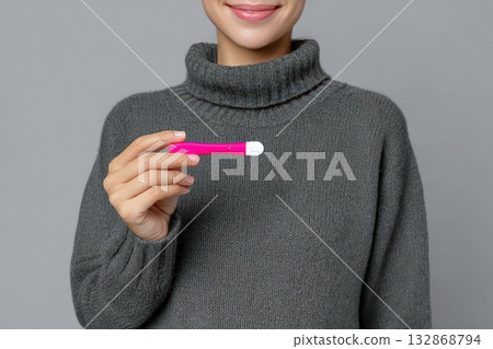 Woman holding a pink pregnancy test with a smile in a cozy gray sweater indoors 132868794