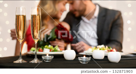 Valentine's Day Celebration. Romantic Couple Having Dinner In Restaurant, Two Champagne Glasses Standing On Table In Foreground, Selective Focus Valentine's Day Celebration. Romantic Couple Having Dinner In Restaurant, Two Champagne Glasses Standing On Table In Foreground, Selective Focus 132871903