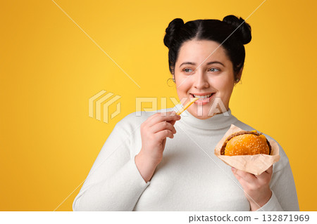 Brunette plump girl eating tasty hamburger and french fries, enjoying fast food and looking aside at copy space over yellow background, panorama 132871969